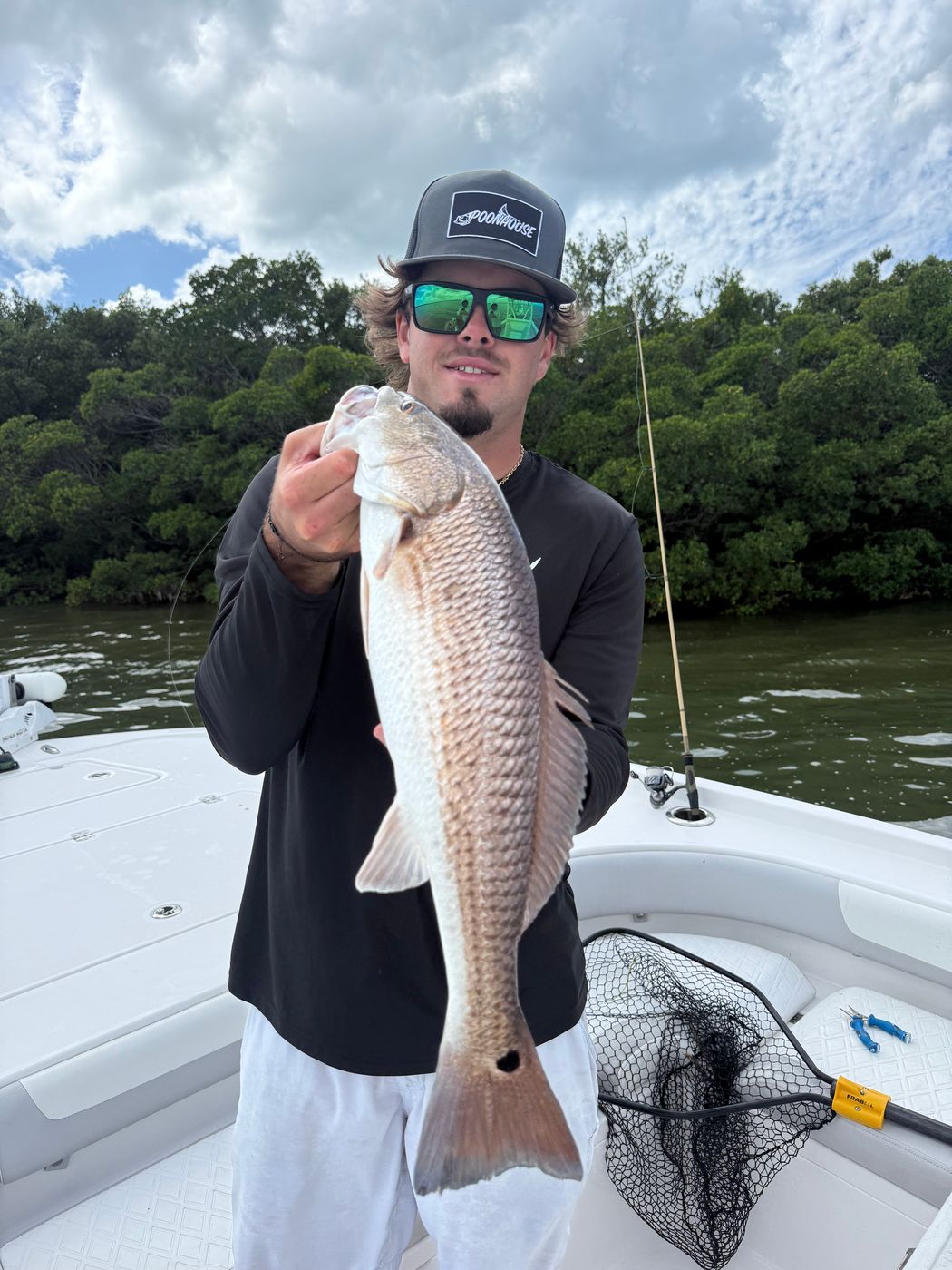 An angler holding a large redfish upright on the boat, a green shoreline and cloudy sky behind