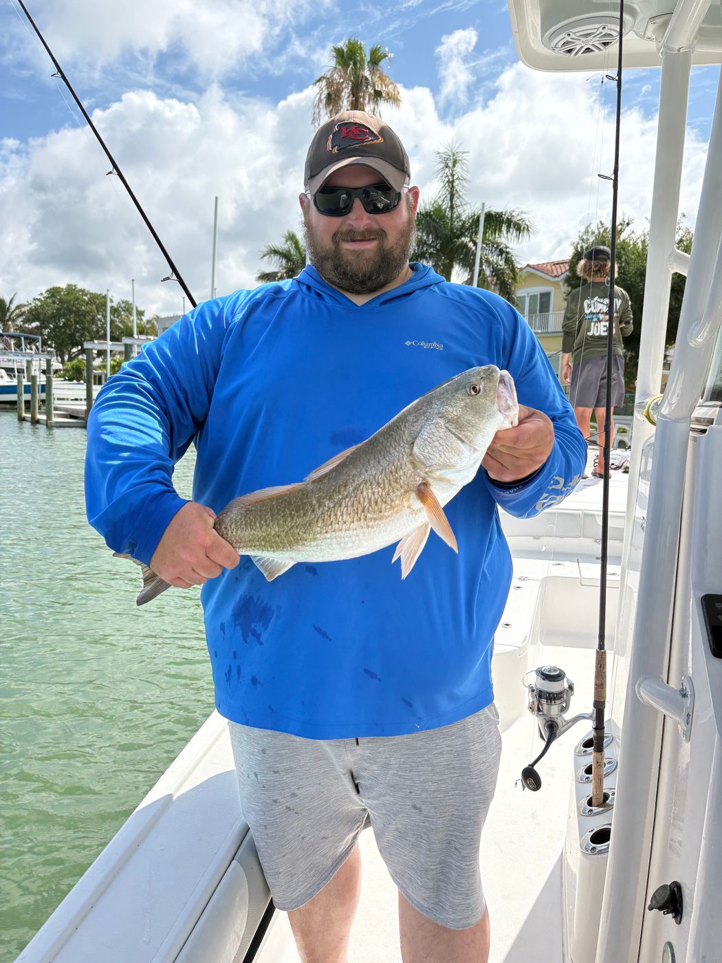 An angler in a blue hooded shirt holding a redfish on the boat, a marina and palms behind him