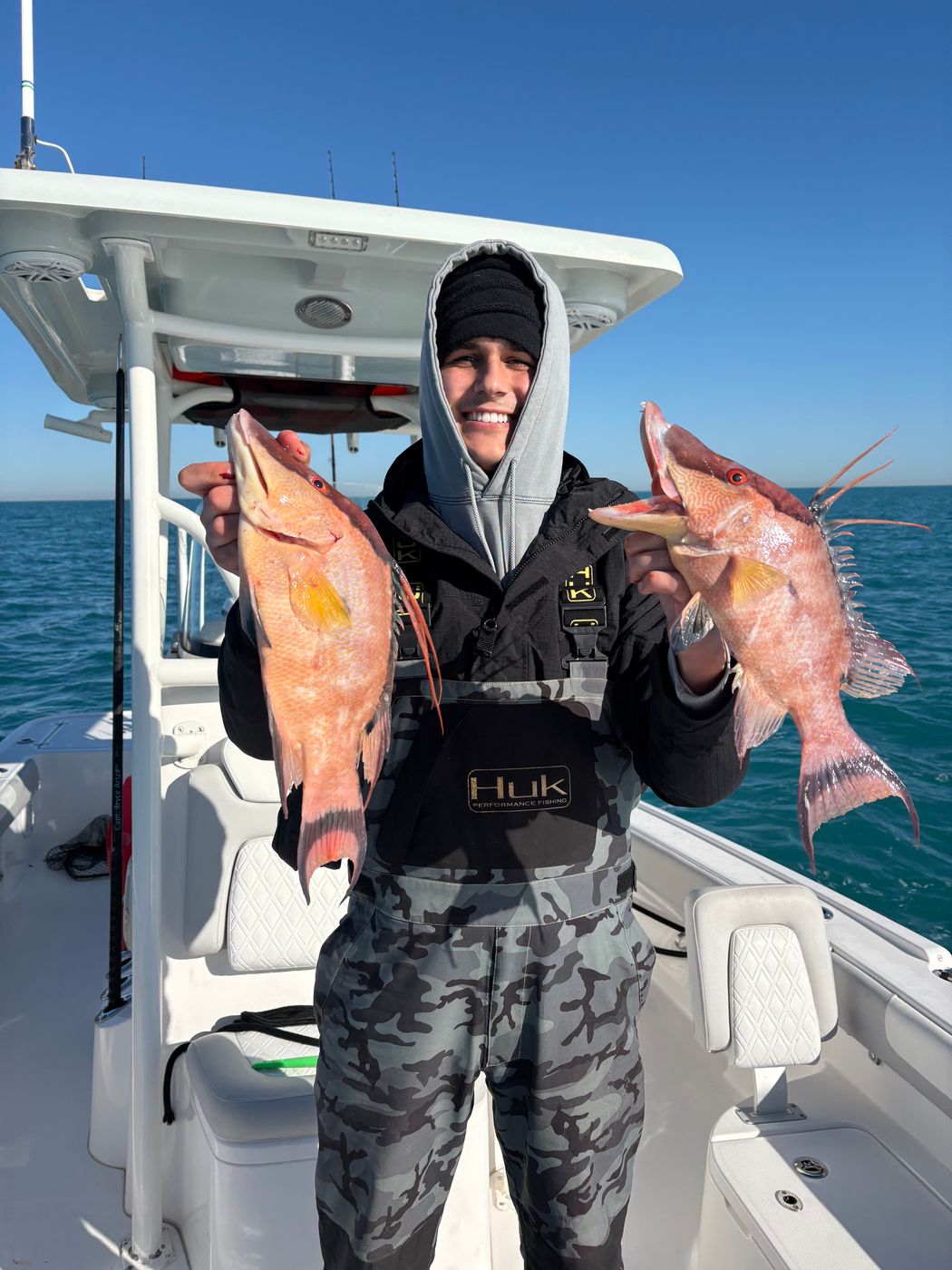 An angler in a hooded jacket holding up two orange-red fish on the boat, the T-top overhead