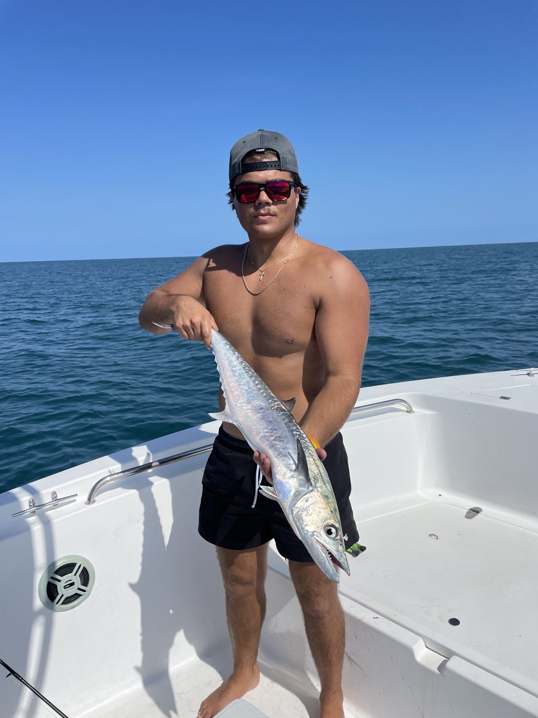 A shirtless angler on the bow holding a long silver fish, blue sky and open water behind