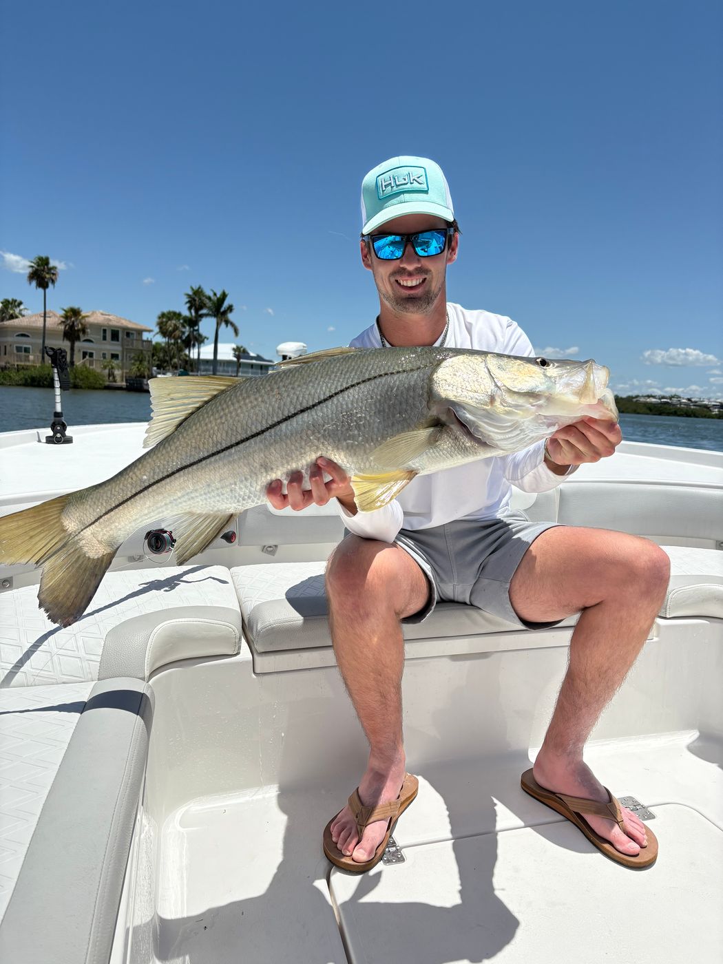 An angler seated on the bow holding a large snook, palms and waterfront homes behind