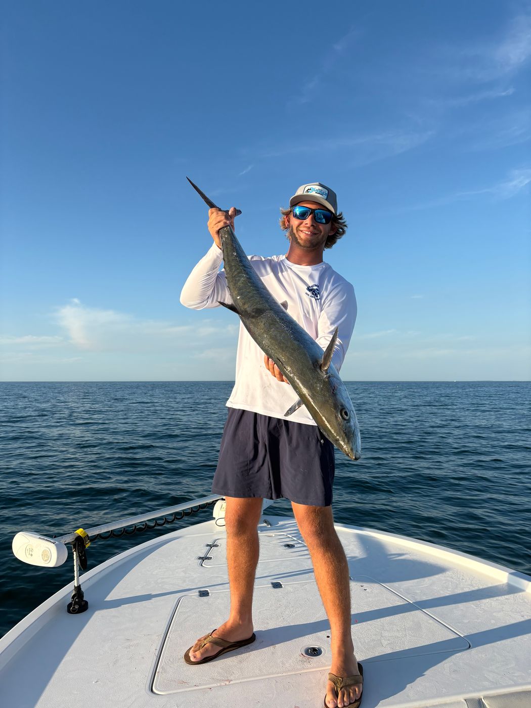 An angler standing on the bow holding up a long fish, open water and blue sky behind