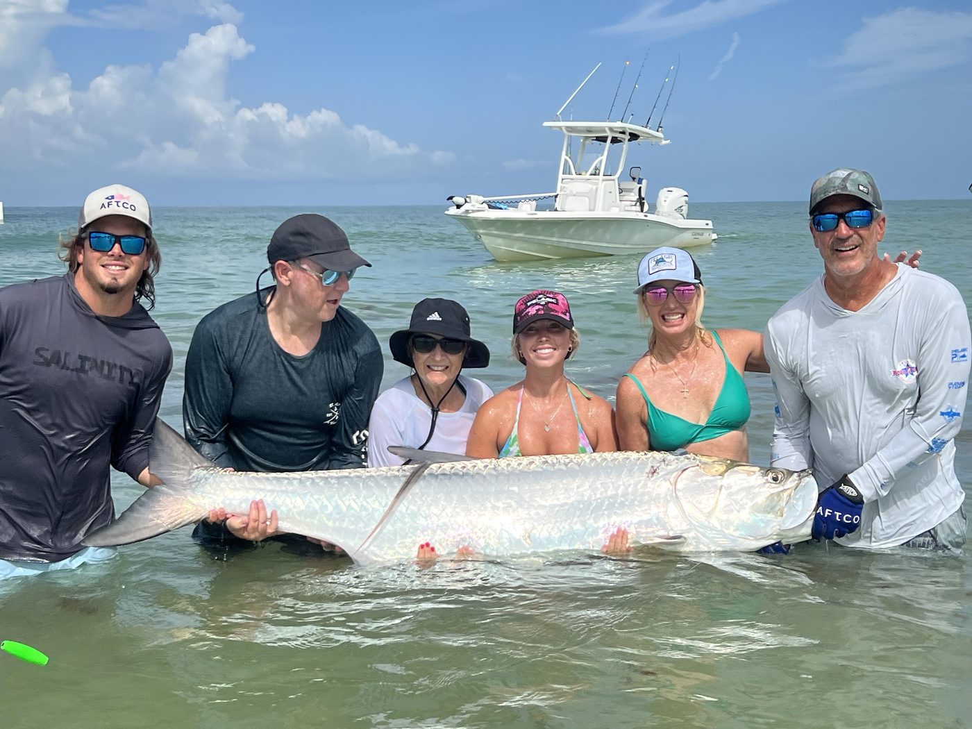 A group of anglers crouched in shin-deep water holding a giant tarpon for a photo, the boat anchored behind