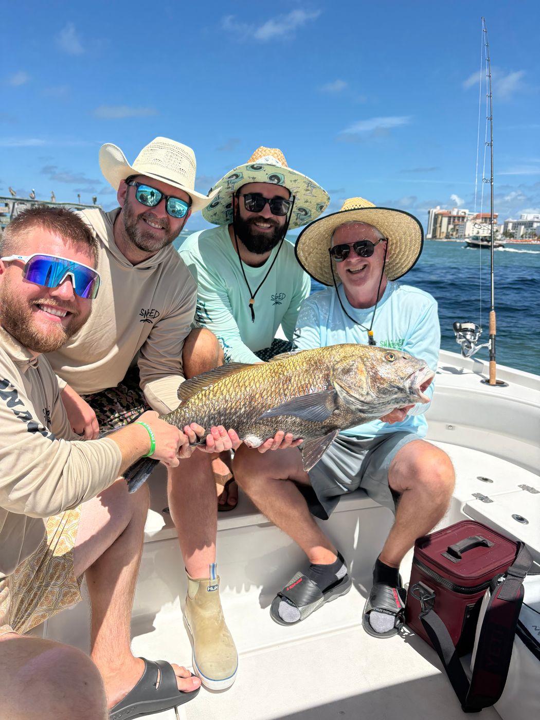 Four anglers in sun hats and sunglasses grinning together over a large fish on the boat, a rod and water behind them