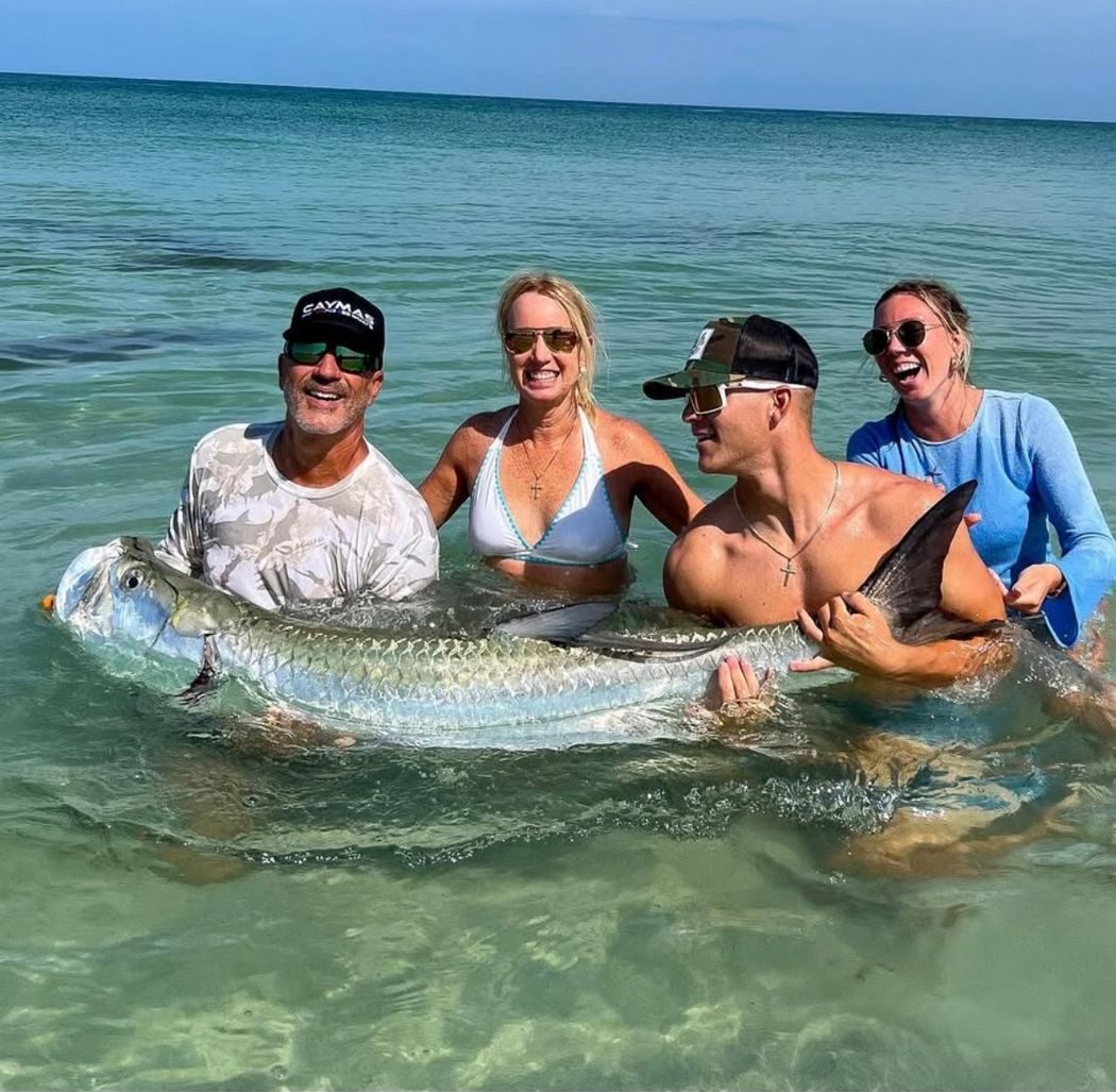 Four people standing in clear, shallow water holding a large tarpon between them, all smiling