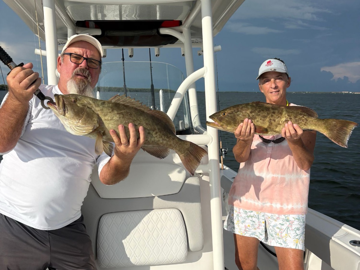 A couple on the boat under the T-top each holding a gag grouper, the bay behind them