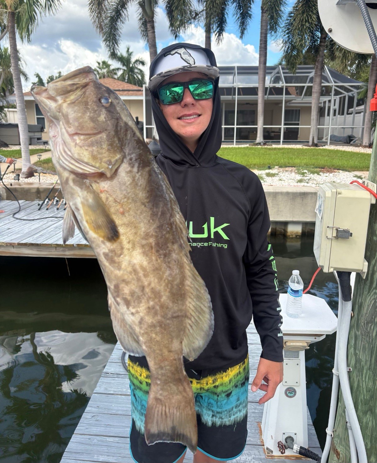 A young angler in a hooded shirt holding a large gag grouper at the dock, palms and waterfront homes behind