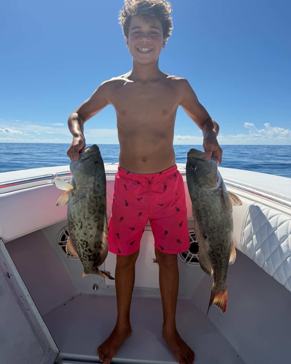 A grinning kid holding two gag grouper on the bow with open water behind