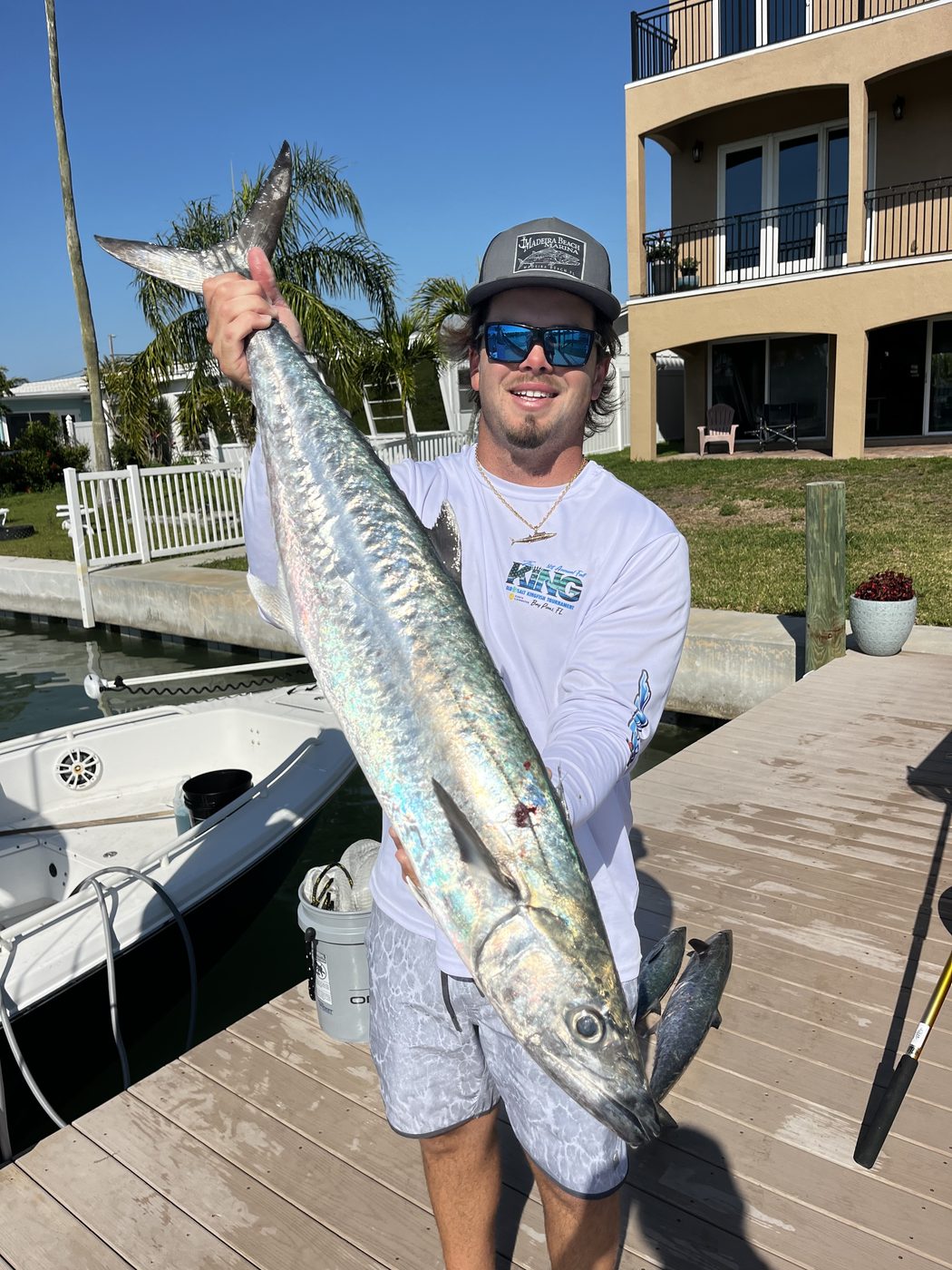 An angler on the dock holding up a long king mackerel, waterfront homes behind