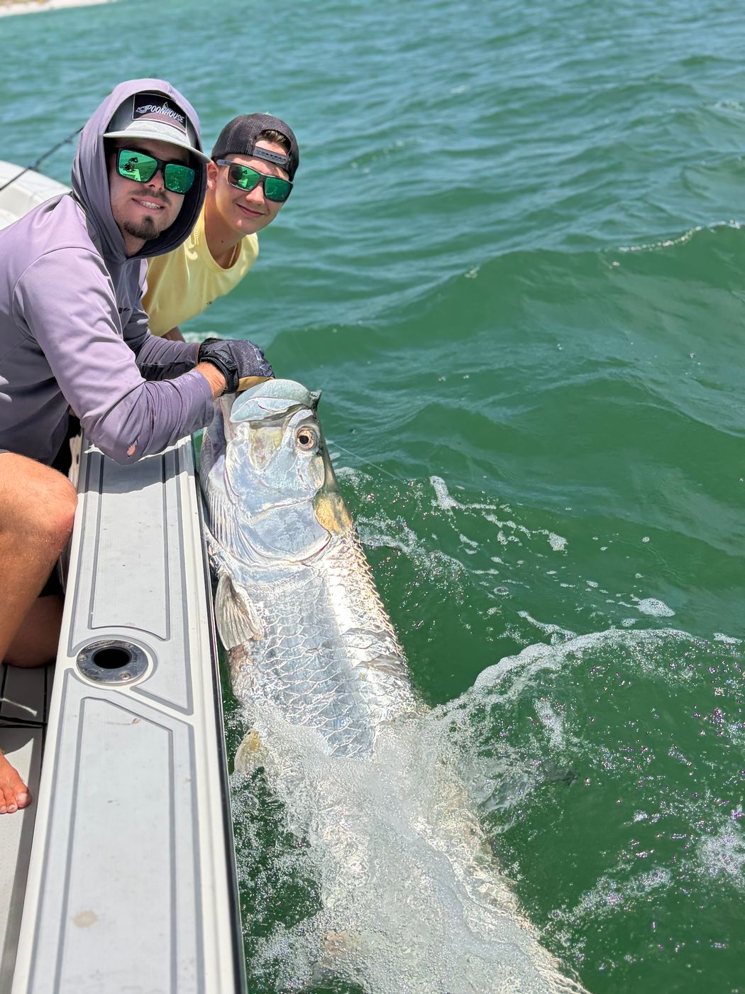 Two anglers leaning over the gunwale as a tarpon thrashes alongside the boat in green water