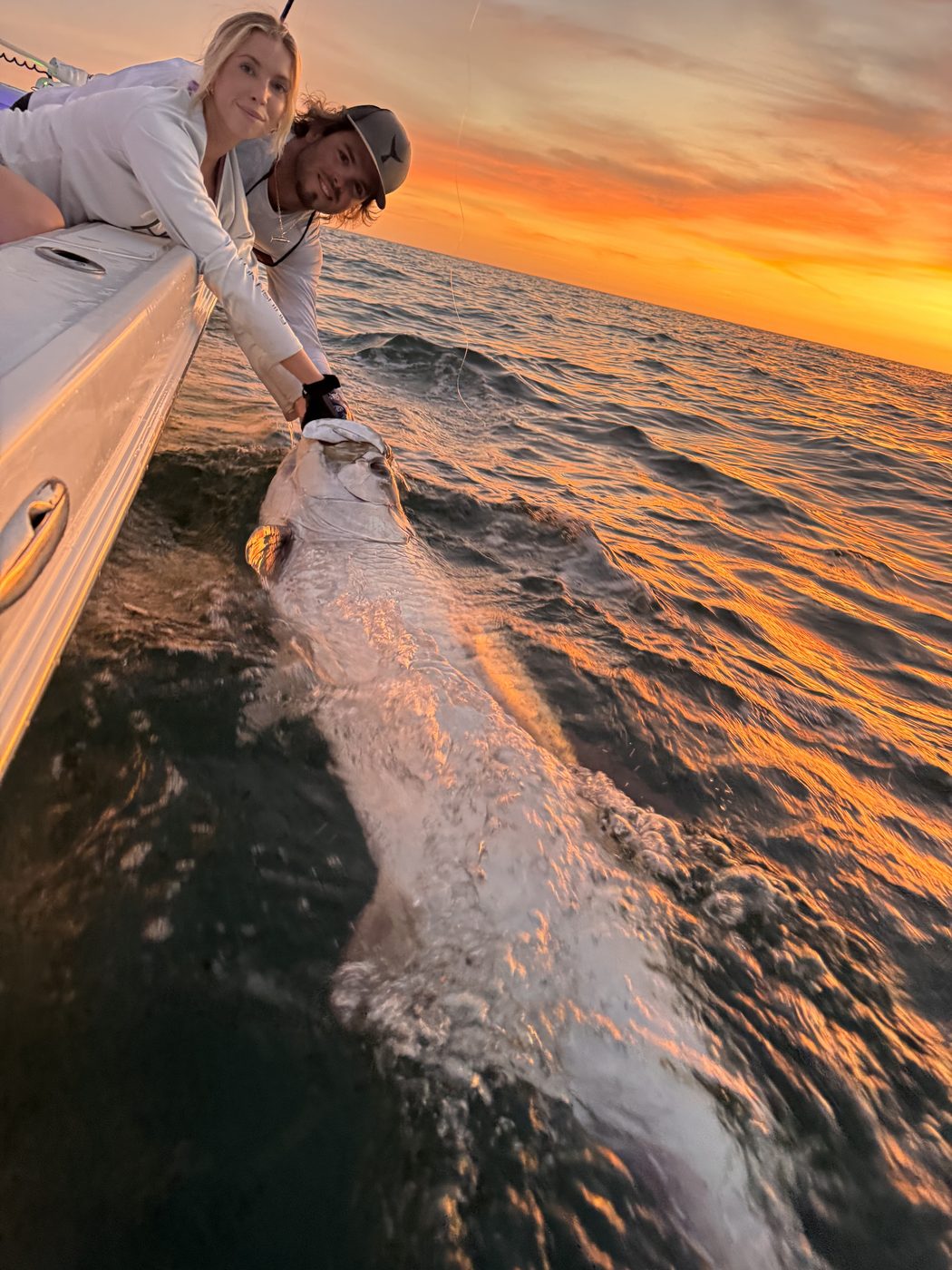 An angler reaching over the gunwale to release a tarpon against an orange sunset