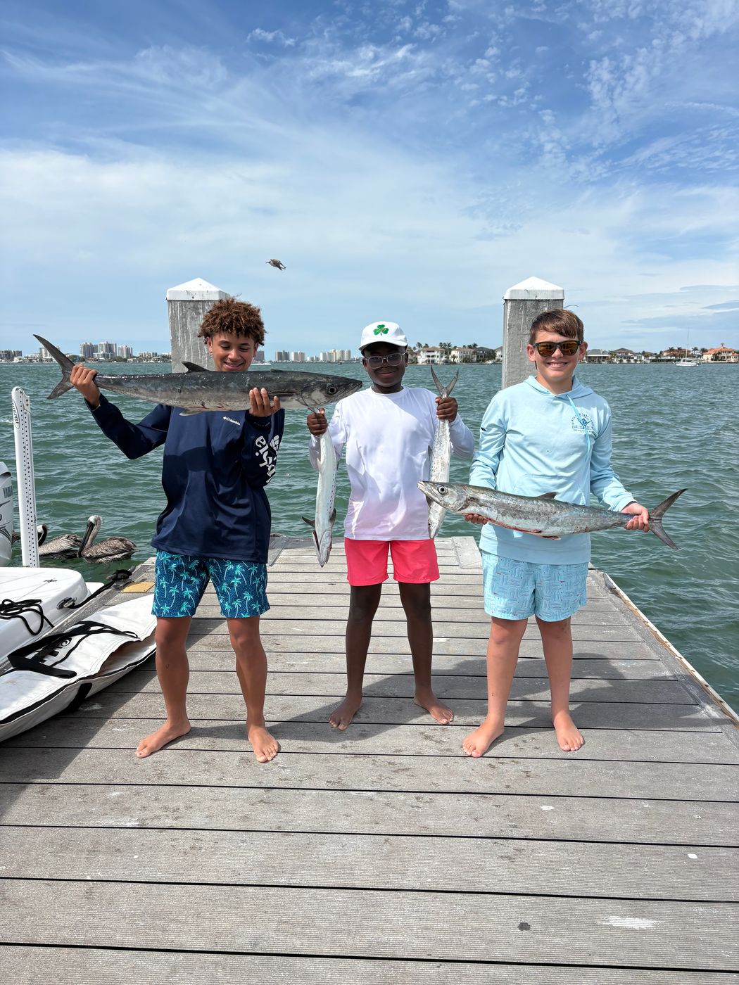 Three kids on a dock each holding a long fish, calm water and a distant shoreline behind