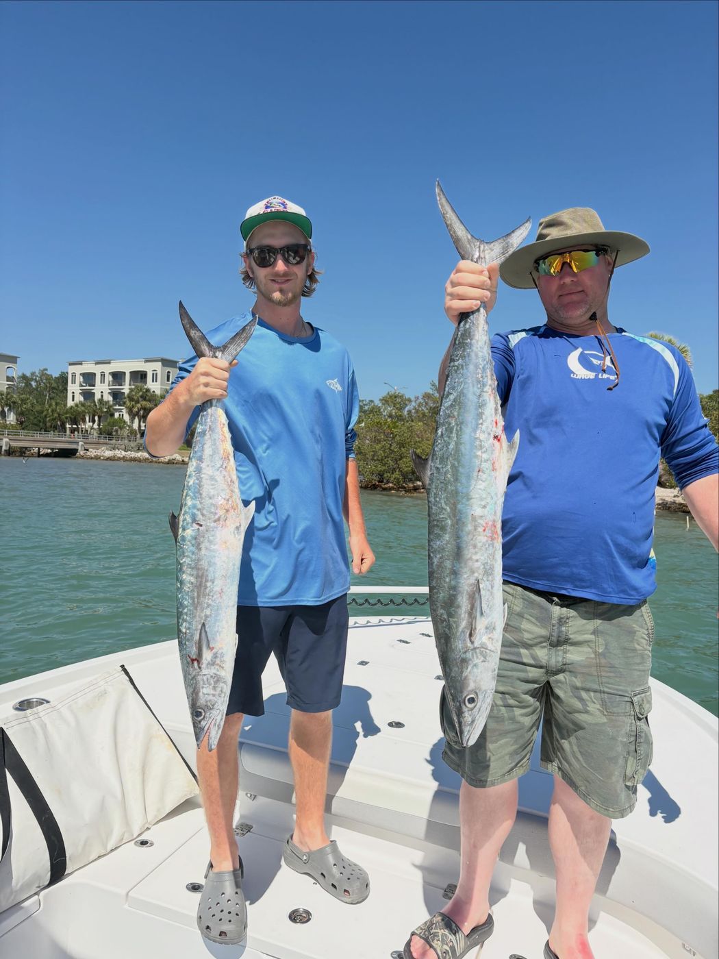 Two anglers on the bow of the boat each holding a long fish, waterfront homes behind
