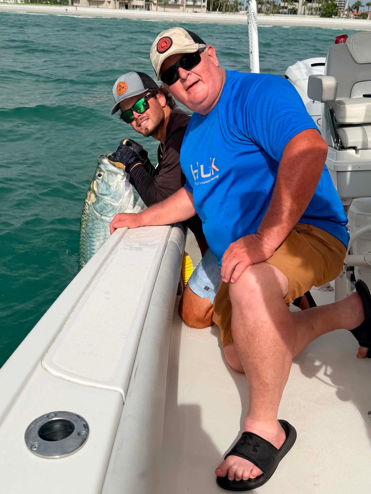 Two anglers at the boat's gunwale with a tarpon alongside, the beach and condos behind