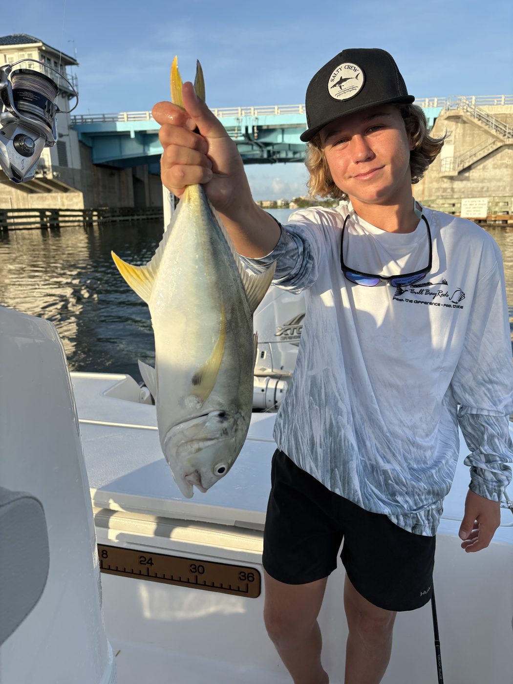 A young angler in a cap holding up a large silvery-yellow fish, a bridge and waterway behind