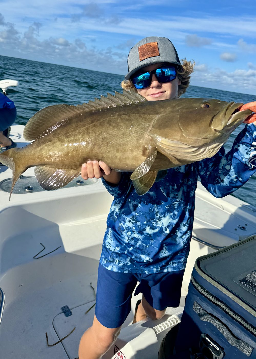 A young angler in a patterned shirt holding a large grouper up to the camera on the boat
