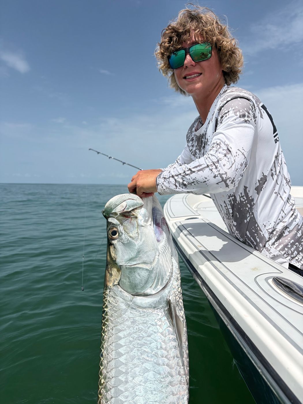 A teenage angler in sunglasses holding a large tarpon at the side of the boat